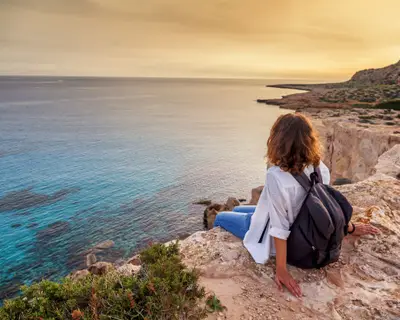 A stylish young woman traveler watches a beautiful sunset on the rocks on the beach, Cyprus, Cape Greco, a popular destination for summer travel in Europe